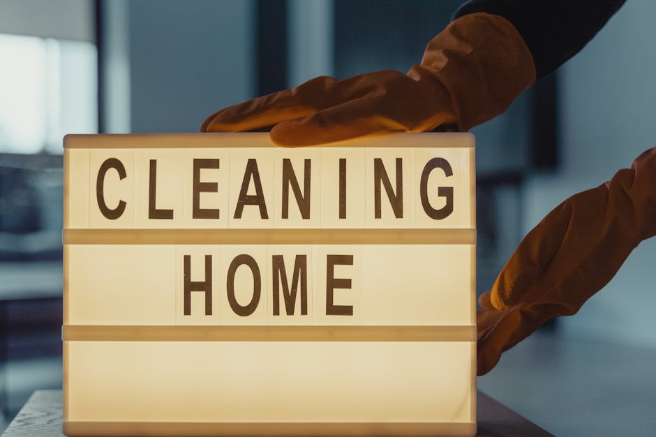 A female cleaning professional from Edgware Carpet Cleaners is indoors, smiling while holding a spray bottle in her right hand and a blue cleaning cloth in her left hand. She is dressed in a bright ye
