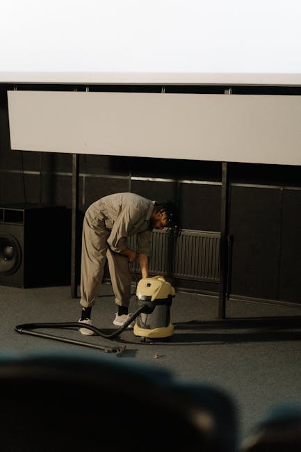 A cleaner, dressed in a light-colored jumpsuit, is operating a vacuum or extraction cleaning machine on a dark carpeted floor in a room with black paneled walls and a black radiator. The room has a white ceiling with a large white panel or screen mounted above. The floor appears to be under deep cleaning or sanitisation, with the equipment suggesting professional surface cleaning. Visible tools include the vacuum unit connected to a hose, and the space is illuminated with soft lighting, emphasizing the cleanliness process conducted by Edgware Carpet Cleaners in a commercial or residential setting near the Broadwalk Centre. The setting demonstrates thorough surface cleaning procedures aimed at maintaining hygiene and freshness of indoor carpets or floors.