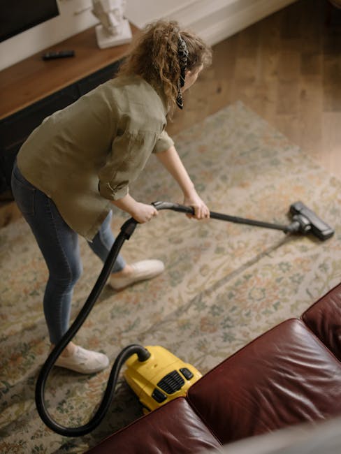 A woman with curly hair, dressed in a light green jacket and blue jeans, is engaged in deep surface cleaning using a vacuum cleaner on a decorative, patterned carpet in a residential living room. The room features wooden flooring along the edges, a brown leather sofa partially visible in the foreground, and a wooden sideboard with a white decorative item on top. Soft, warm lighting illuminates the area, highlighting the cleanliness of the carpet and the tidy, well-maintained appearance of the room. The scene illustrates professional domestic cleaning practices, with Edgware Carpet Cleaners providing thorough surface sanitisation and deep cleaning of carpets in a home environment.